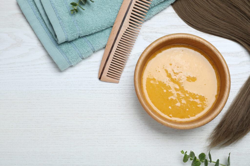 Bol de masque capillaire maison jaune doré, peigne en bois, serviette et mèche de cheveux sur une table en bois blanc