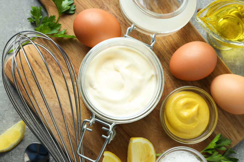 Bocal de mayonnaise maison entouré d'œufs, d'un fouet, d'un ramequin de moutarde, d'huile et de quartiers de citron