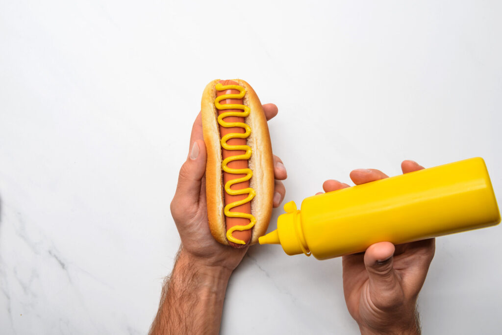 Mains qui pressent un flacon de moutarde jaune au-dessus d'un hot-dog, sur un plan de travail en marbre blanc