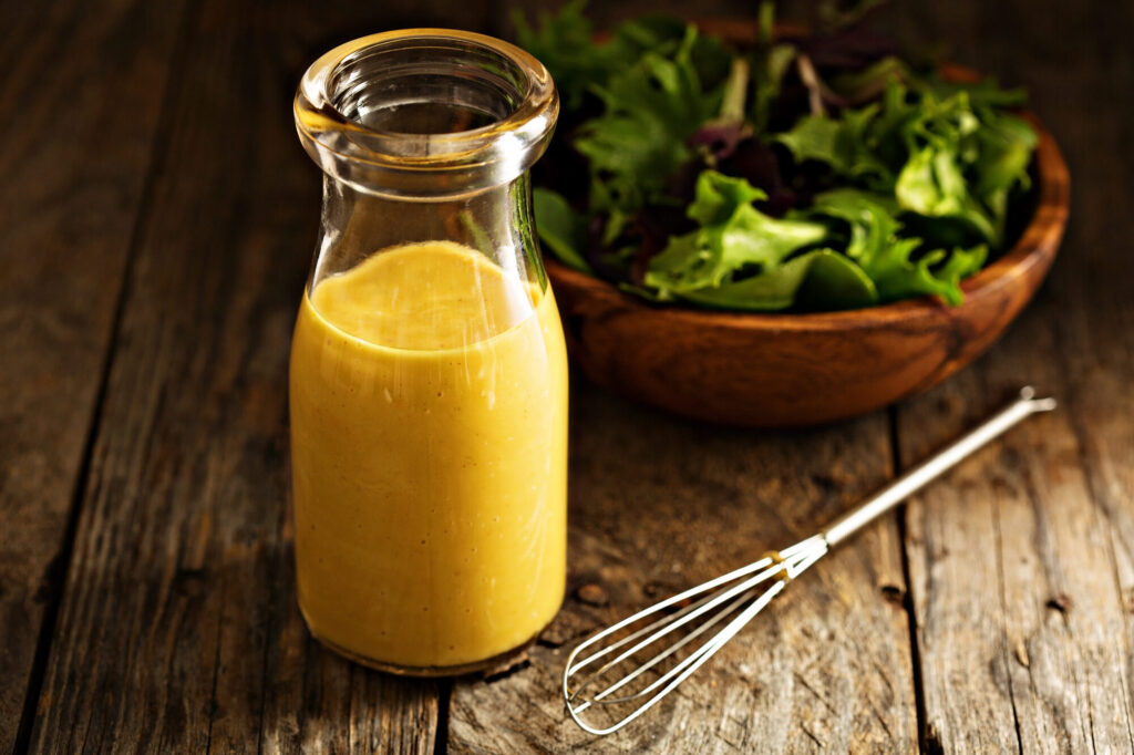 Bouteille de vinaigrette à la moutarde jaune doré, fouet et saladier de jeunes pousses sur une table en bois