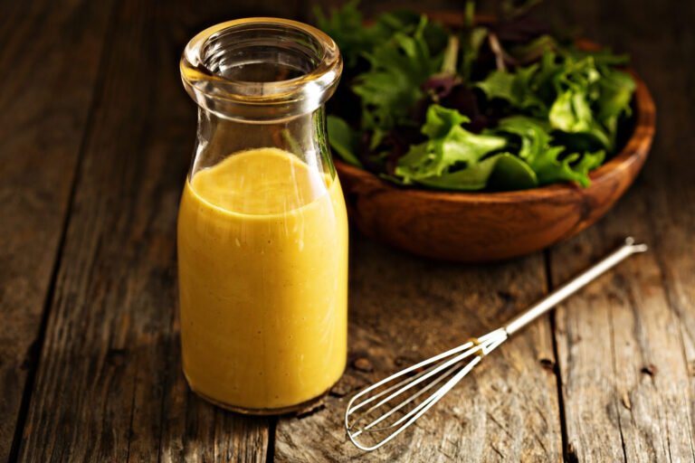 Bouteille de vinaigrette à la moutarde jaune doré, fouet et saladier de jeunes pousses sur une table en bois