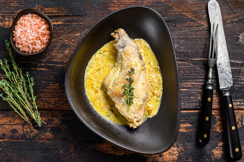 Cuisse de lapin dans une sauce jaune crémeuse, servie dans une assiette noire, avec du thym, du sel rose et des couverts sur une table en bois