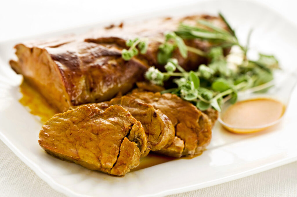 Filet de porc doré, tranché en médaillons sur un plat blanc, avec une coupelle de sauce et des herbes fraîches