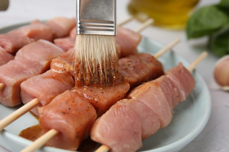 Pinceau qui étale une marinade sur des brochettes de viande crue posées sur une assiette