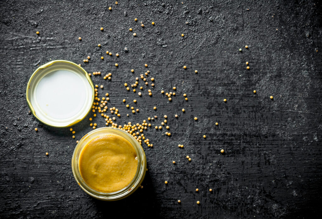 Petit bocal de moutarde jaune vif ouvert, son couvercle à côté, et graines de moutarde éparpillées sur un fond noir