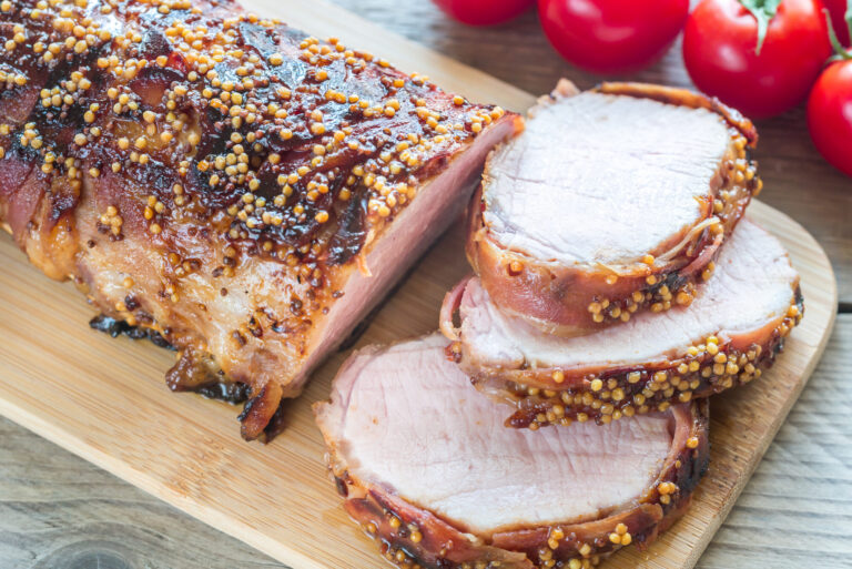 Rôti de porc bardé de lard et couvert de graines de moutarde, tranché sur une planche, avec des tomates cerises
