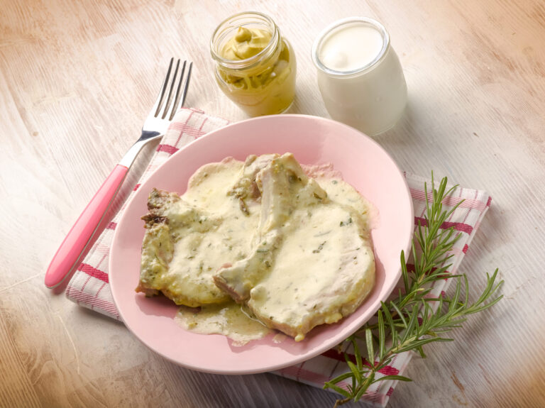 Deux tranches de viande nappées d'une sauce crémeuse à la moutarde sur une assiette rose, avec un pot de moutarde et du romarin