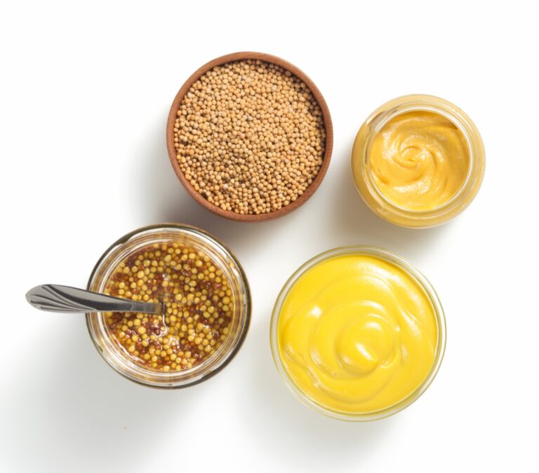 Quatre moutardes et un bol de graines vus de dessus sur fond blanc : graines entières, moutarde lisse, moutarde à l'ancienne en bocal et moutarde jaune vif