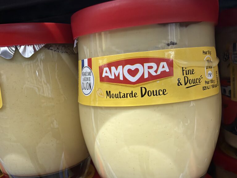 Pots de moutarde douce Amora en verre, à couvercle rouge et étiquette jaune, sur une étagère de magasin